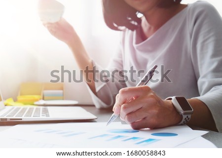 Asian woman work at home or internet cafe  by holding pen to writing information to prepare data for the presentation, sale report for meeting, shopping online, chat and hold cup of coffee for break  Royalty-Free Stock Photo #1680058843