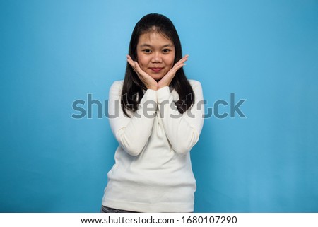 Portrait of Young beautiful asian women using white T-shirt with blue isolated background, look at the camera and smile, both hand on the chin.