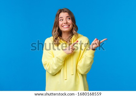 Cheerful hipster girl in yellow hoodie, with blond short haircut, pointing and looking right side with pleased happy smile, promote product or company services, stand blue background