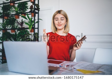 Content female business owner in red casual t shirt closing eyes and crossing fingers while waiting answer from partner on mobile phone at stylish office 