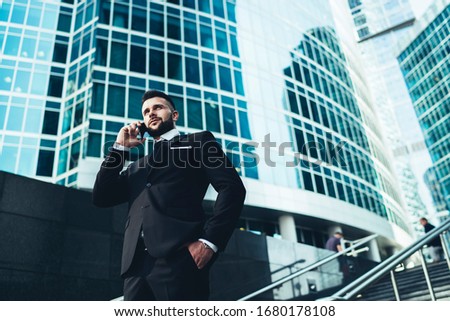 Serious and pensive man in suit talking on phone while standing on stairs against  background of large buildings