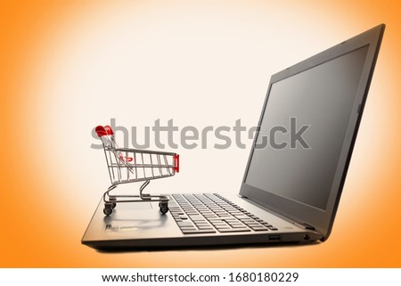 A laptop with a toy shopping cart sitting on the keyboard. Isolate on an orange background. Copy space. Side view. Concept of online shopping and modern technologies