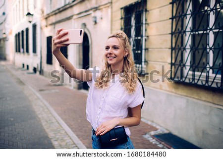 Peaceful slender woman with blond hair in white blouse shooting selfie on footpath on old narrow street on blurred background