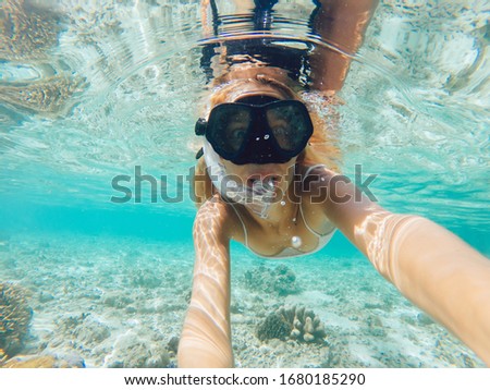 Young beautiful woman with blonde hair in swimsuit and snorkeling mask swimming under surface of water and taking selfie near sea bottom with coral reef