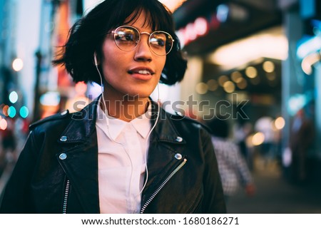 Millennial woman in fashionable clothing listening audio book during evening walk in New York City, youthful female enjoying adolescent music podcast during nightlife lifestyle in Manhattan Royalty-Free Stock Photo #1680186271