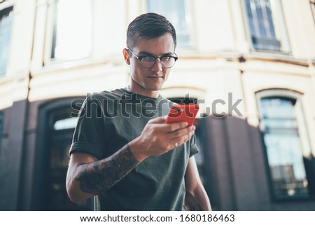 Millennial hipster guy in spectacles sharing photos to social network using 4g wireless for browsing internet and posting publications via smartphone application, young man checking time on cellular