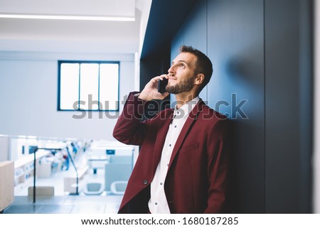 Focused looking up young bearded male in white shirt holding red jacket in hand calling on mobile phone in modern creative workplace leaning on wall