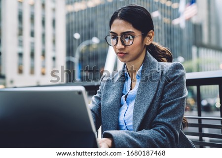 Confident ethnic young businesswoman in elegant outfit and eyeglasses sitting at table outdoors browsing tablet on terrace of New York city 