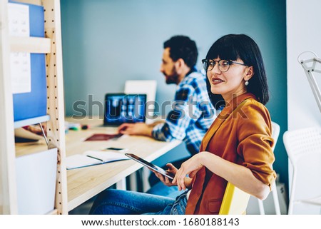 Portrait of confident millennial woman in eyewear holding tablet and sitting nets to male colleague during working process, young attractive female employee using technology for planning on meeting