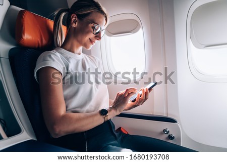 Female tourist in classic eyewear checking received email in social networks during internet messaging on board in jetliner, Caucasian woman booking return tickets connected to high speed wifi Royalty-Free Stock Photo #1680193708