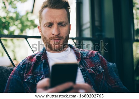 Crop pensive bearded male in casual clothes focusing on screen and interacting with smartphone while sitting on soft couch against blurred metallic fence and green trees