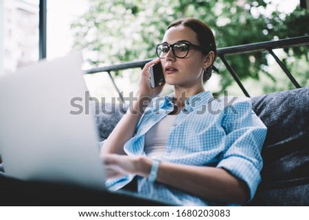 From below pretty pensive adult female in glasses making phone call while sitting on sofa at laptop on balcony against blurred green trees in daylight