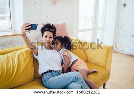 Smiling African American young casual mother taking selfie with curly upset child while hugging on yellow home couch in light room