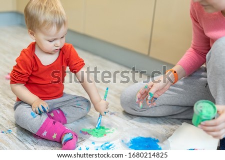 Adorable cute caucasian little blond toddler boy enjoy having fun finger painting with young beautiful mother at home indoors . Cheerful happy kids smiling drawing masterpiece art picture