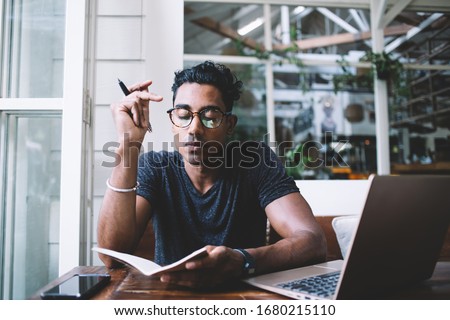 Concentrated serious Hispanic man in eyeglasses thinking and taking notes while sitting at table with laptop and smartphone in tropical cafe Royalty-Free Stock Photo #1680215110