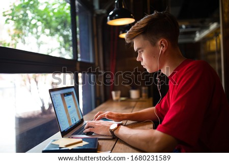 Hipster guy in casual wear listening to music via headphones and online learning via laptop computer while relaxing in coffee shop. Man skilled blog writer typing text on notebook gadget 