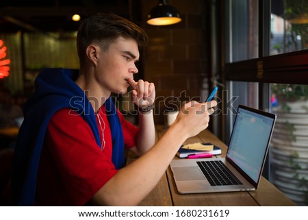 Young hipster guy with thoughtful look checking e-mail on mobile phone while sitting with laptop computer in coffee shop. Man using apps on cell telephone 