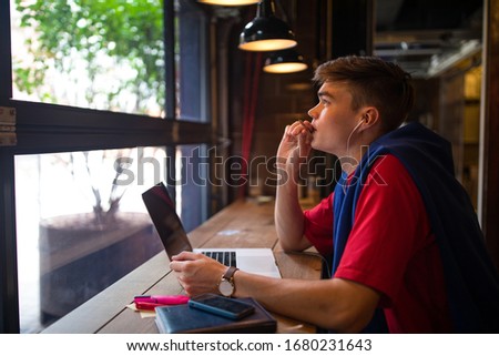 Young pondering man thinking about new ideas while sitting with laptop computer in coffee shop interior. Pensive male student having online education via notebook gadget 