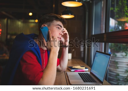 Young handsome man in trendy casual wear having cellphone conversation and using laptop computer for online education while relaxing in coffee shop 