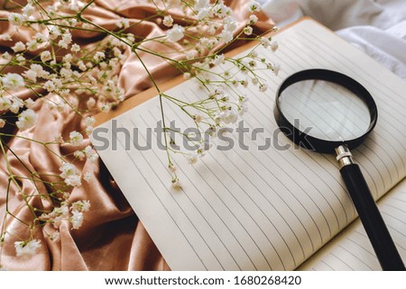 Spring composition, white gypsophila flowers with notebook and magnifying glass on the gold satin fabric