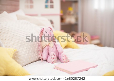 Handmade plush rabbit made of fabric sits on a cozy bed in the children 's room. Nursery there is a Dollhouse and a lot of toys. On the bed there are a lot of soft pillows stars. copy space.