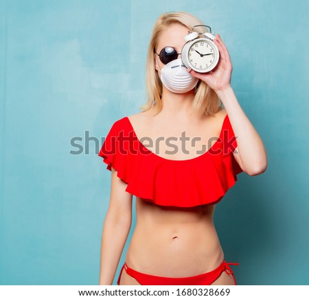 Blonde woman in bikini and face mask with alarm clock on blue background 