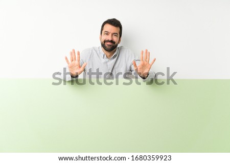 Young handsome man with beard holding a big green empty placard counting ten with fingers