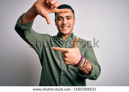 Young brazilian man wearing casual shirt standing over isolated white background smiling making frame with hands and fingers with happy face. Creativity and photography concept.