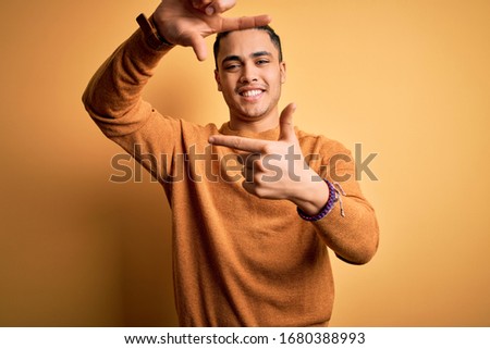 Young brazilian man wearing casual sweater standing over isolated yellow background smiling making frame with hands and fingers with happy face. Creativity and photography concept.