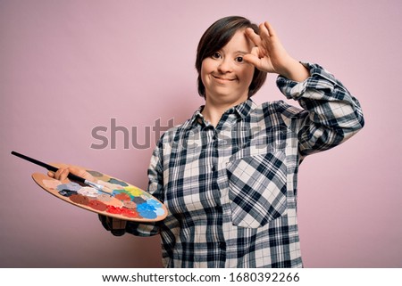 Young down syndrome artist woman holding painter palette and paintbrush over pink background doing ok sign with fingers, excellent symbol