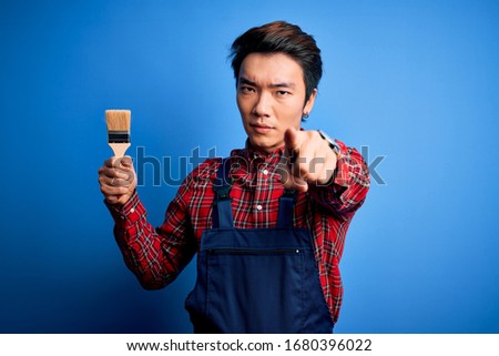 Young handsome chinese painter man wearing apron painting using brush pointing with finger to the camera and to you, hand sign, positive and confident gesture from the front