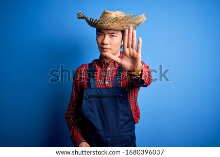 Young handsome chinese farmer man wearing apron and straw hat over blue background doing stop sing with palm of the hand. Warning expression with negative and serious gesture on the face.