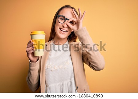 Young beautiful businesswoman drinking cup of takeaway coffee over yellow background with happy face smiling doing ok sign with hand on eye looking through fingers