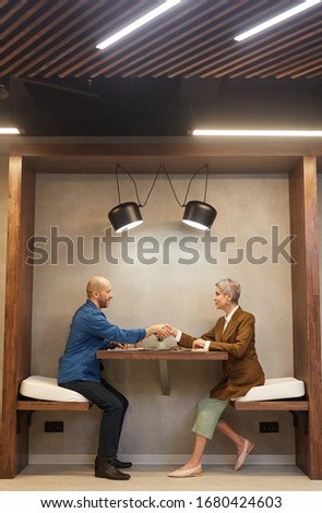 Side view full length portrait of two people shaking hands while sitting at table in indoor cafe during business meeting, copy space