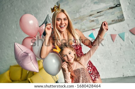 Happy birthday! Mother and daughter having fun together and holding paper crowns, pretty mom and lovely cute daughter wears similar festive dress and birthday hats. Family lovely moments.