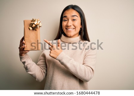 Young beautiful asian woman holding birthday gift standing over isolated white background very happy pointing with hand and finger