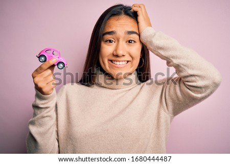 Young beautiful asian woman holding pink small car standing over isolated background stressed with hand on head, shocked with shame and surprise face, angry and frustrated. Fear and upset for mistake.
