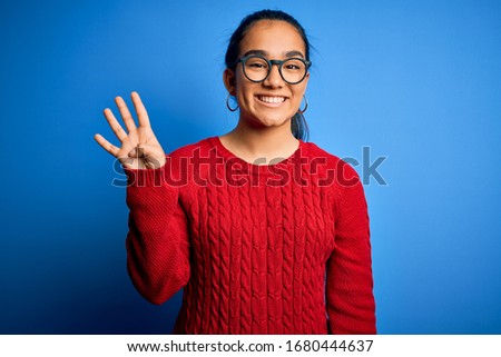 Young beautiful asian woman wearing casual sweater and glasses over blue background showing and pointing up with fingers number four while smiling confident and happy.