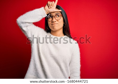 Young beautiful asian woman wearing casual sweater and glasses over red background making fun of people with fingers on forehead doing loser gesture mocking and insulting.