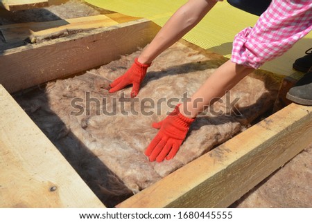 Constructing a rooftop a building contractor in protective gloves is lying glass wool batt insulation between the roofbeams for energy efficiency of the house attic insulation. Royalty-Free Stock Photo #1680445555