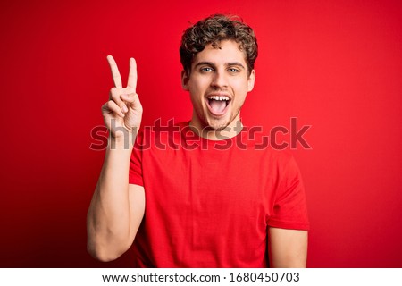 Young blond handsome man with curly hair wearing casual t-shirt over red background smiling with happy face winking at the camera doing victory sign with fingers. Number two.