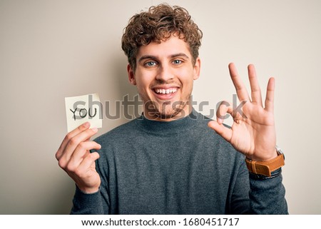 Young blond man with curly hair holding paper with you message over white background doing ok sign with fingers, excellent symbol