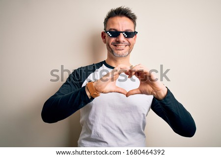 Young handsome man wearing funny thug life sunglasses meme over white background smiling in love showing heart symbol and shape with hands. Romantic concept.