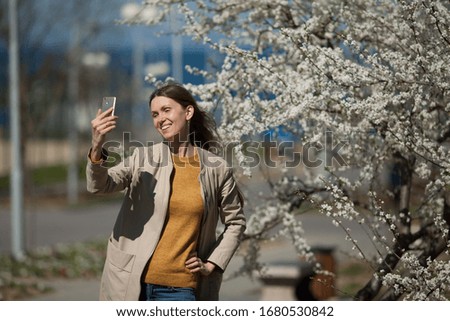A young woman takes a selfie on her smartphone against the background of a luxuriously blooming tree in spring. The concept of lush blooming, travel, technology.