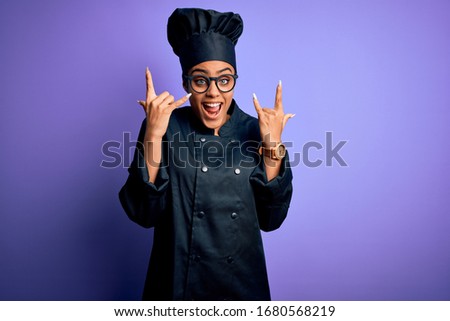 Young african american chef girl wearing cooker uniform and hat over purple background shouting with crazy expression doing rock symbol with hands up. Music star. Heavy music concept.