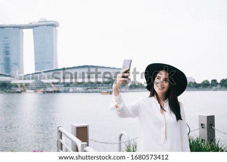 Stylish touristic woman with smile standing at pier and taking selfie on mobile phone on background of designed architectural building and river 