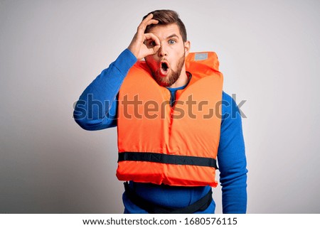 Young blond tourist man with beard and blue eyes wearing lifejacket over white background doing ok gesture shocked with surprised face, eye looking through fingers. Unbelieving expression.