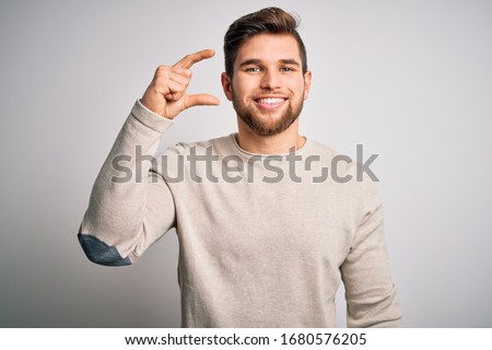 Young handsome blond man with beard and blue eyes wearing casual sweater smiling and confident gesturing with hand doing small size sign with fingers looking and the camera. Measure concept.