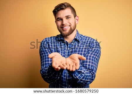 Young blond businessman with beard and blue eyes wearing shirt over yellow background Smiling with hands palms together receiving or giving gesture. Hold and protection