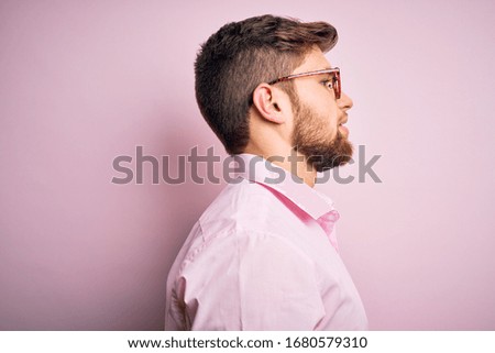 Young handsome blond man with beard and blue eyes wearing pink shirt and glasses looking to side, relax profile pose with natural face with confident smile.